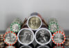 US Mint Coin Production Slows as America 250 Rollout Expands Close-up view of cylindrical coin rolls with US coins peeking out from orange and green wrappers, stacked together inside a container.