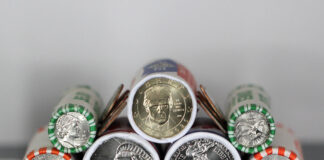 US Mint Coin Production Slows as America 250 Rollout Expands Close-up view of cylindrical coin rolls with US coins peeking out from orange and green wrappers, stacked together inside a container.