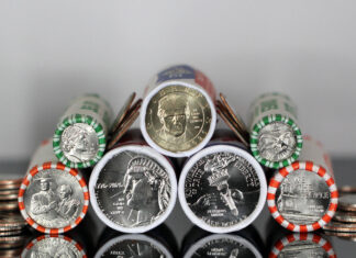 US Mint Coin Production Slows as America 250 Rollout Expands Close-up view of cylindrical coin rolls with US coins peeking out from orange and green wrappers, stacked together inside a container.