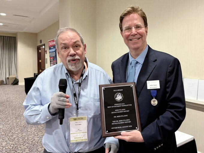 Dr. Michael Fuljenz stands at left holding an award plaque and addressing seated guests while Scott A. Travers, at right, presents the plaque at the Numismatic Literary Guild awards luncheon.